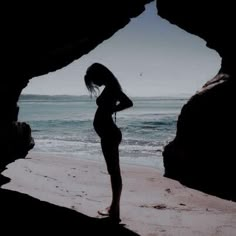 a woman standing in front of a cave on top of a beach next to the ocean