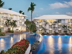 an outdoor swimming pool surrounded by palm trees and buildings with lights on at night time