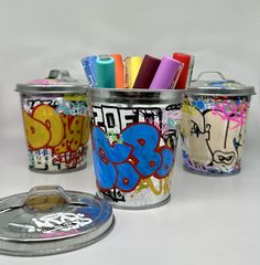 three tins filled with markers and pens on top of a white table next to a coin