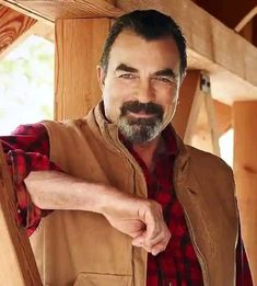 a man with a goatee and plaid shirt standing in front of a wooden structure