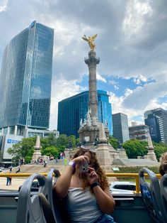 a woman sitting on top of a bus next to a tall building with a statue in the background