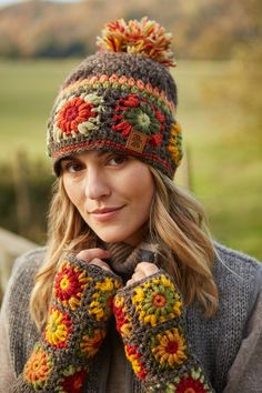 a woman wearing a knitted hat and mittens with flowers on the top, standing in front of a field