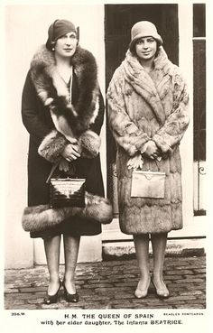 two women standing next to each other in front of a building wearing fur coats and hats