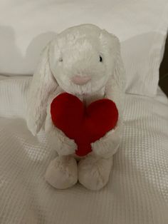 a white stuffed rabbit holding a red heart on top of a bed with white sheets