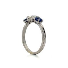 This exquisite three-stone engagement ring features a .93 carat antique cushion-cut diamond with VS2 clarity and J color. Flanked by two vibrant round blue sapphires, each weighing .46 carats, the sapphires add elegance to the overall design. The polished platinum band showcases intricate milgrain detailing. Decorative baskets cradle each gemstone, ensuring beauty from every angle. This ring blends refined craftsmanship with eye-catching details, making it a stunning choice. Luxury Three-stone Sapphire Promise Ring, Luxury Three Stone Sapphire Promise Ring, Classic Three Stone Jewelry With Lab-created Sapphire, Sapphire Three Stone Round Cut Diamond Ring, Three Stone Sapphire Diamond Ring With Round Cut, Three Stone Lab-created Sapphire Diamond Ring, Fine Jewelry Three Stone Lab-created Sapphire Ring, Timeless Three-stone Sapphire Diamond Ring, Timeless Sapphire Three-stone Diamond Ring