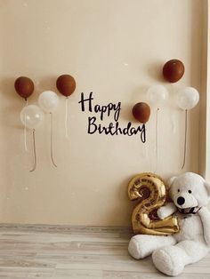 a white teddy bear sitting next to balloons and a happy birthday sign on the wall