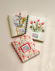 three embroidered notebooks with flowers on them are sitting next to each other and one has a notepad in front of it