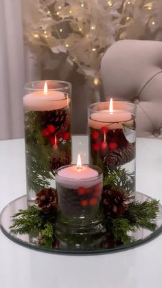 three lit candles sitting on top of a glass plate covered in greenery and pine cones