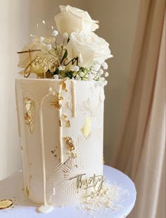 a white cake with gold decorations and flowers on top is sitting on a table in front of a window