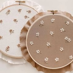 two embroidered hoops with flowers on them sitting next to each other, one in white and the other in beige