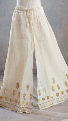 PRODUCT DETAILS This is a beautiful mul-mul cotton Off-White Palazzo Pants with Gota patti borders and gold leather flowers on the Shararaa.. It is beautifully made in soft mul-mul cotton fabric and has a A-Line Flary hem. It also has an attached lining inside. It has a stretchy elastic band and a tying string for a good fit. The Skirt can fit up to size 40 inch waist and can be tightened with the strings for narrower waist. Size & Fit One Size Waist: 28 (expandable to 42) Height: 39.5 inches In White Palazzo Pants Outfit Indian, White Palazzo Pants Outfit, Palazzo Pants Outfit Indian, Pallazo Suit, Palazzo Pants Design, Women Sharara, Indian Pants, Skirt Lehenga, Palazzo Pants Outfit