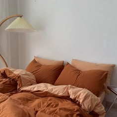 an unmade bed with orange sheets and pillows in a white room next to a lamp