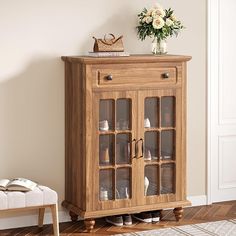 a small wooden cabinet with glass doors and flowers on top, in front of a white wall