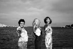 three women standing next to each other in front of the ocean pointing at something with their fingers