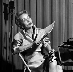 a woman sitting in a chair reading a piece of paper next to a piano and microphone