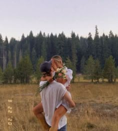 a man carrying a woman in the middle of a field with pine trees behind him