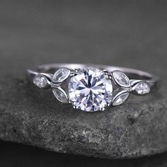 a white gold engagement ring with pear shaped diamonds and leaves on the band, sitting on top of a rock