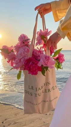 a person holding a bag with flowers in it on the beach near the ocean at sunset