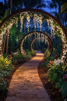 an archway with flowers and lights on it in the middle of a garden at night