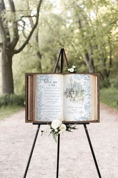 an open book with flowers on it sitting on top of a easel in the middle of a dirt road