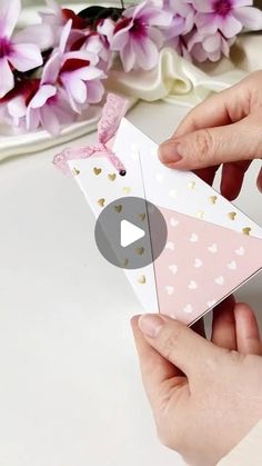 two hands are holding an origami triangle with hearts on it and pink flowers in the background