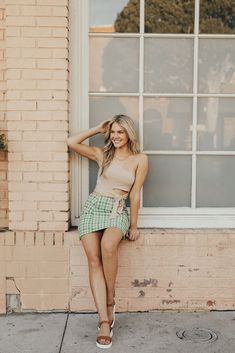 a woman leaning up against a window in front of a brick building wearing a tan top and green checkered shorts