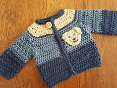 a crocheted baby sweater with a teddy bear on the front and bottom, sitting on a wooden surface