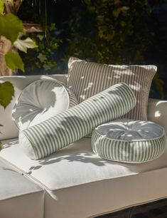 pillows and cushions are sitting on a white couch in the sun outside with greenery behind them