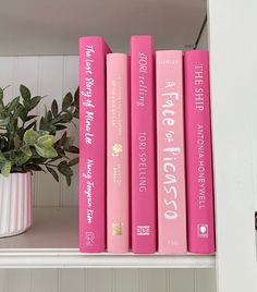 three pink books sitting on top of a white shelf next to a potted plant