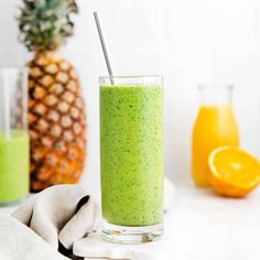 a glass filled with green smoothie next to two oranges and a pineapple