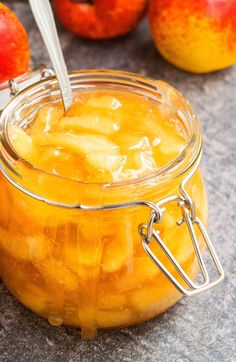 a glass jar filled with apple jam next to apples on a counter top and in the background, there is a spoon sticking out of it