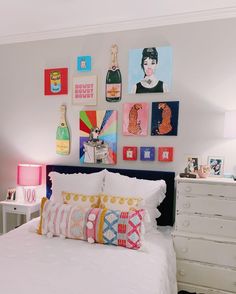 a white bed topped with lots of pillows next to a wall covered in pictures and paintings