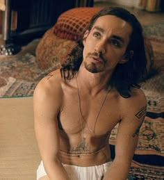 a shirtless man sitting on the floor with his hands in his pockets and looking off to the side