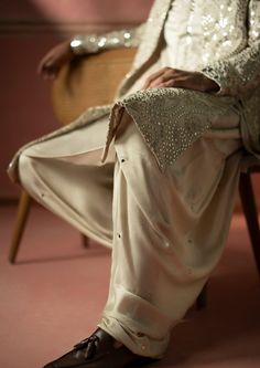 a man sitting on top of a wooden chair wearing white pants and a jacket with sequins