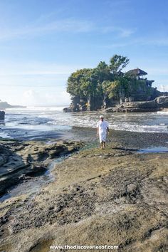 a man standing on top of a rock covered beach next to the ocean with an island in the background