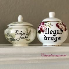 two porcelain jars sitting on top of a white shelf with words painted on the lids