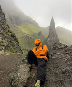 a man in an orange jacket sitting on top of a rocky hill next to mountains