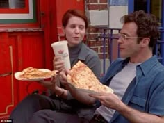 two people eating pizza and drinking coffee on the street in front of a red building