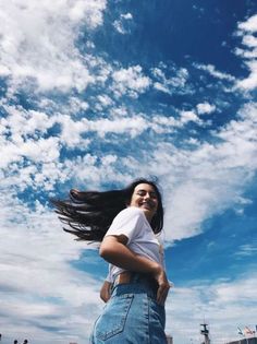 a woman with long hair standing in front of a blue sky and white fluffy clouds