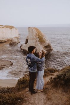 a man and woman standing on a path next to the ocean with cliffs in the background