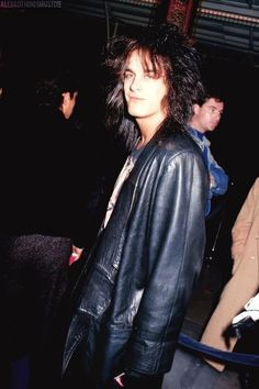 a man with black hair and leather jacket standing in front of a group of people