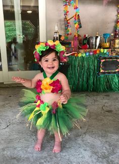 a baby girl wearing a hula skirt and flower headband smiling at the camera