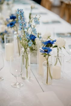 blue and white flowers in glass vases on a table with wine glasses, candles and napkins