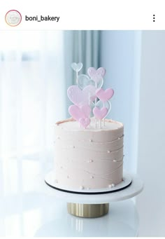 a pink cake with hearts on top sitting on a white table next to a window