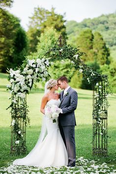 a bride and groom standing in front of an arch with white flowers on the grass