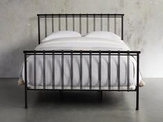 an iron bed frame with white linens and pillows on it, against a concrete wall