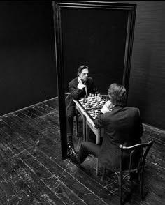 two men sitting at a table playing chess in front of a mirror with the reflection of them