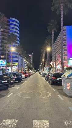 cars parked on the street in front of tall buildings and palm trees at night time