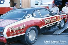 two men standing next to a red and white car with the word plymouth on it