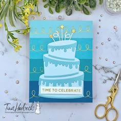 a birthday card with a blue cake on it and gold scissors next to it, surrounded by greenery